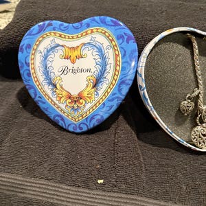 Puede incluir: Una caja de hojalata en forma de coraz&oacute;n con un dise&ntilde;o floral azul y amarillo. La tapa de la caja tiene la palabra "Brighton" escrita en letra cursiva. La caja est&aacute; abierta y el interior est&aacute; vac&iacute;o.