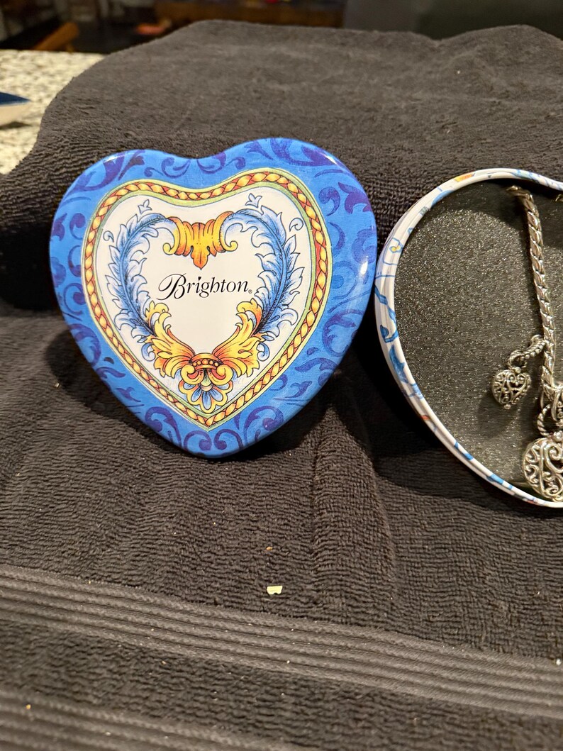 Puede incluir: Una caja de hojalata en forma de coraz&oacute;n con un dise&ntilde;o floral azul y amarillo. La tapa de la caja tiene la palabra "Brighton" escrita en letra cursiva. La caja est&aacute; abierta y el interior est&aacute; vac&iacute;o.