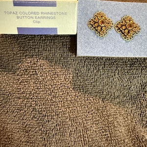 May include: A pair of gold-toned clip-on earrings with topaz coloured rhinestones. The earrings are in a box that says "Topaz Coloured Rhinestone Button Earrings Clip".