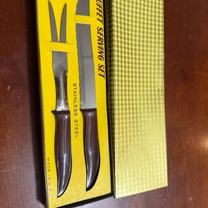 May include: A buffet serving set in a yellow box. The set includes a carving knife and a serving fork with brown handles. The box is labeled "BUFFET SERVING SET" and "STAINLESS STEEL". The set is made in the U.S.