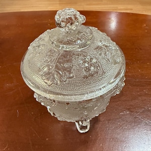 May include: A clear glass candy dish with a lid. The dish has a round shape with a decorative grape design and sits on three small feet. The lid has a decorative knob on top.
