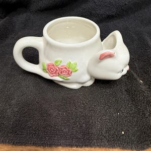May include: White ceramic cat-shaped mug with a handle. The mug features a cat's head and body, with pink accents on the ears and nose. Pink roses and green leaves decorate the side of the mug. The mug is on a dark surface.