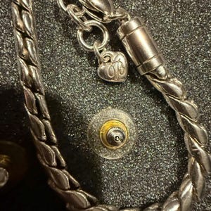 Puede incluir: Un brazalete de cadena de plata con un dije de coraz&oacute;n y un cierre magn&eacute;tico. El brazalete est&aacute; trenzado y tiene un acabado plateado.