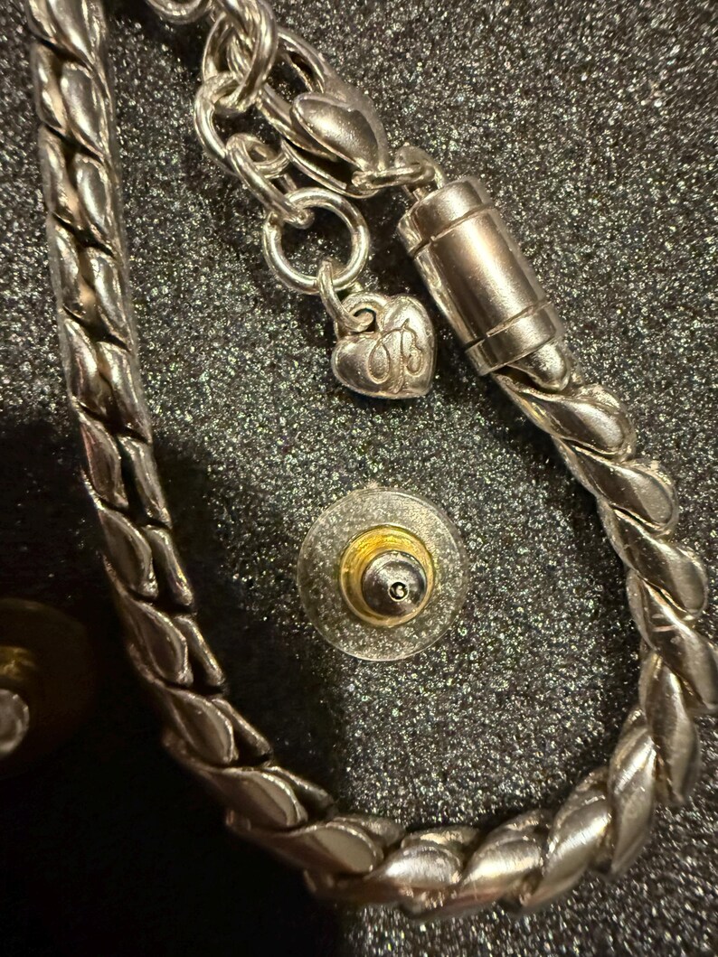 Puede incluir: Un brazalete de cadena de plata con un dije de coraz&oacute;n y un cierre magn&eacute;tico. El brazalete est&aacute; trenzado y tiene un acabado plateado.