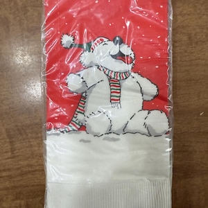 May include: A pack of red and white paper napkins featuring a cartoon polar bear wearing a green and red hat and a striped scarf. The bear is sitting in the snow against a red background.