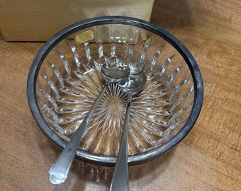 May include: A clear glass serving bowl with a silver rim and two silver serving spoons. The bowl has a starburst design in the center and a ribbed pattern around the sides. The spoons are resting inside the bowl.