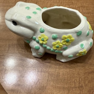May include: White ceramic frog planter with green and yellow floral accents. The frog has a wide open mouth, forming the planter.