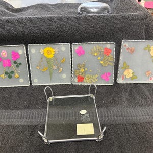 May include: Four square glass coasters with pressed flowers and leaves. The coasters are arranged in a row on a dark gray surface. A metal coaster holder is in the foreground.
