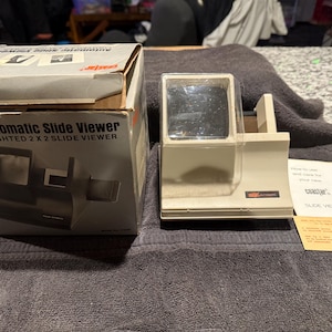 Puede incluir: Un visor de diapositivas automático de plástico blanco con una imagen en blanco y negro de un visor de diapositivas en la caja. La caja dice "Automatic Slide Viewer Lighted 2 x 2 Slide Viewer".