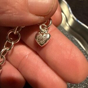Puede incluir: Un brazalete de plata con un dije en forma de coraz&oacute;n. El dije tiene un dise&ntilde;o detallado y est&aacute; unido a una cadena de plata.