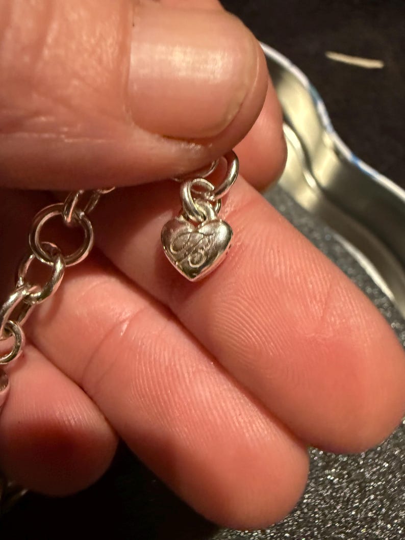 Puede incluir: Un brazalete de plata con un dije en forma de coraz&oacute;n. El dije tiene un dise&ntilde;o detallado y est&aacute; unido a una cadena de plata.