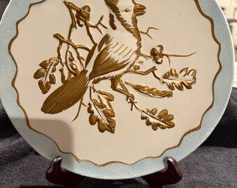 May include: A white ceramic plate with a blue rim and a gold embossed design of a bird perched on a branch with leaves.