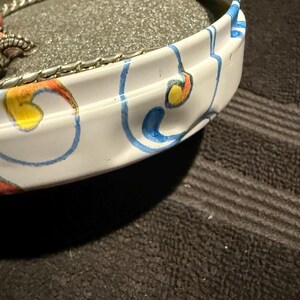 Puede incluir: Una lata de metal blanca con un dise&ntilde;o floral azul y amarillo. La lata tiene un inserto de espuma y se utiliza para guardar joyas.