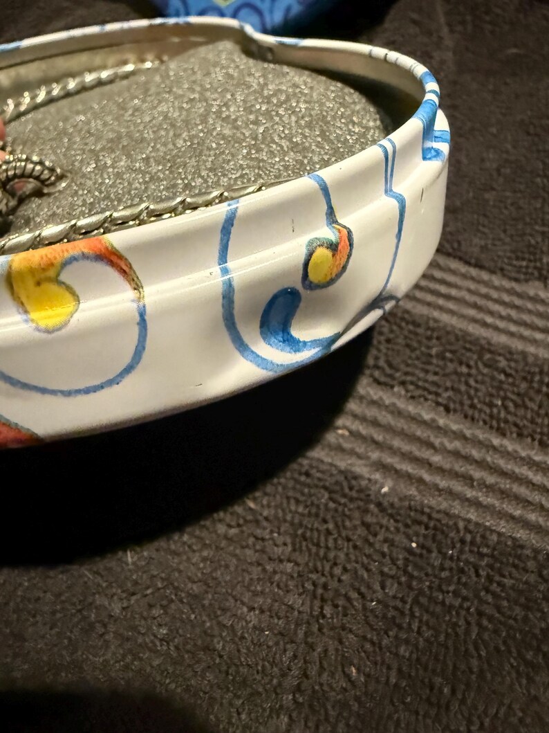 Puede incluir: Una lata de metal blanca con un dise&ntilde;o floral azul y amarillo. La lata tiene un inserto de espuma y se utiliza para guardar joyas.