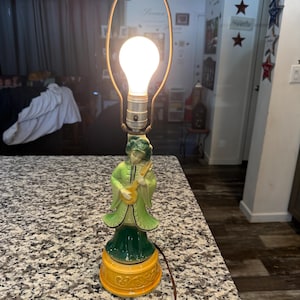 Puede incluir: Una lámpara de mesa de cerámica con una base verde y amarilla con forma de mujer tocando un laúd. La lámpara tiene una bombilla blanca y una pantalla de metal.