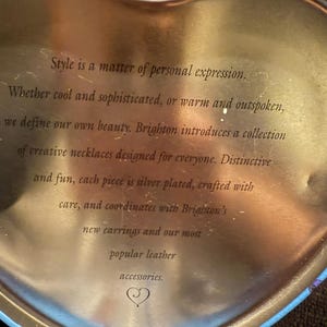 Puede incluir: Una lata dorada en forma de coraz&oacute;n con un borde azul. La lata tiene un mensaje sobre las joyas de Brighton: "El estilo es una cuesti&oacute;n de expresi&oacute;n personal. Ya sea que seas cool y sofisticado, o c&aacute;lido y franco, definimos nuestra propia belleza. Brighton presenta una colecci&oacute;n de collares creativos dise&ntilde;ados para todos. Distintivos y divertidos, cada pieza est&aacute; plateada, elaborada con cuidado y se coordina con los nuevos pendientes de Brighton y nuestros accesorios de cuero m&aacute;s populares."