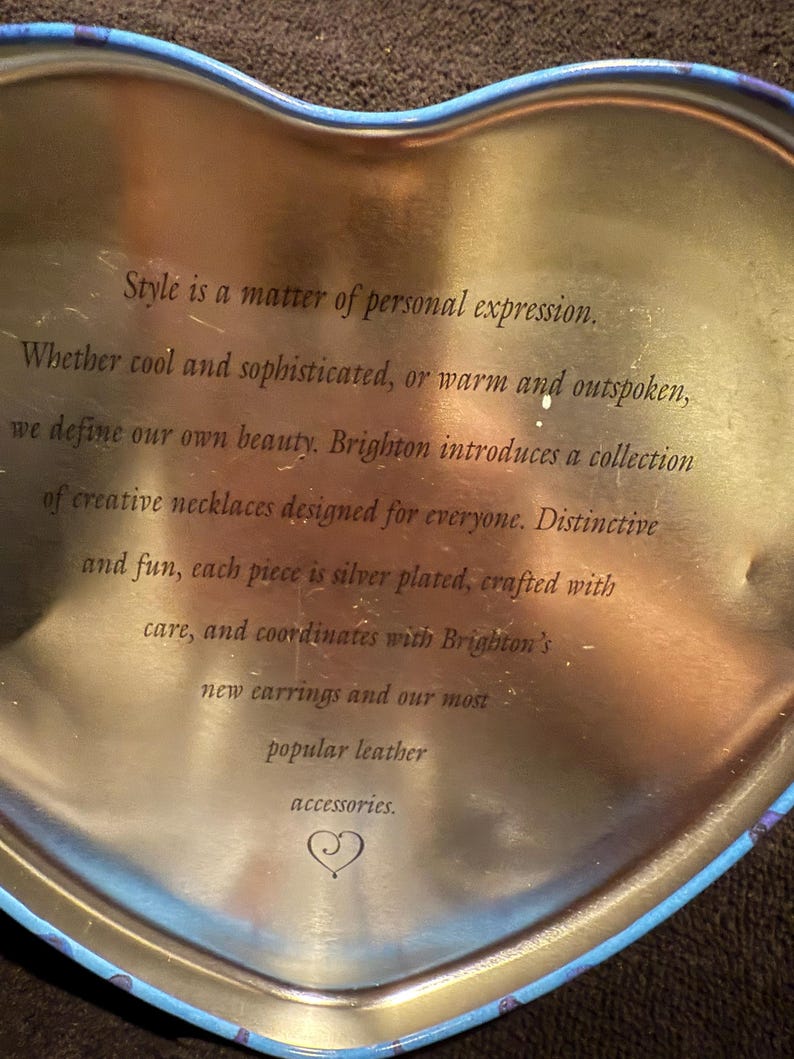 Puede incluir: Una lata dorada en forma de coraz&oacute;n con un borde azul. La lata tiene un mensaje sobre las joyas de Brighton: "El estilo es una cuesti&oacute;n de expresi&oacute;n personal. Ya sea que seas cool y sofisticado, o c&aacute;lido y franco, definimos nuestra propia belleza. Brighton presenta una colecci&oacute;n de collares creativos dise&ntilde;ados para todos. Distintivos y divertidos, cada pieza est&aacute; plateada, elaborada con cuidado y se coordina con los nuevos pendientes de Brighton y nuestros accesorios de cuero m&aacute;s populares."