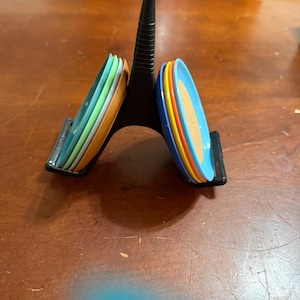 Puede incluir: Un soporte decorativo negro de varios niveles que sostiene una pila de posavasos coloridos. Los posavasos son en tonos de azul, naranja, amarillo, verde y blanco. El soporte está sobre una superficie de madera.