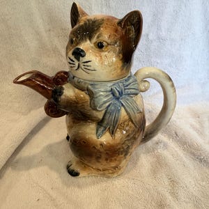 Könnte beinhalten: Eine keramische Teekanne in Katzenform mit braunen, beigen und schwarzen Fellmustern. Die Katze trägt eine blaue Schleife und hält einen braunen Ausguss. Der Griff ist ein geschwungener Schwanz. Ein einzigartiger und skurriler Küchenartikel.