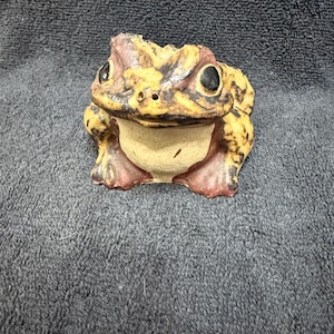 May include: A ceramic frog figurine with brown, yellow, and pink paint. The frog has black eyes and a wide, toothy grin.