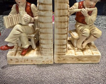 May include: A pair of vintage ceramic bookends featuring a boy playing an accordion and a boy playing a flute. The bookends are painted in shades of cream, brown, and red.