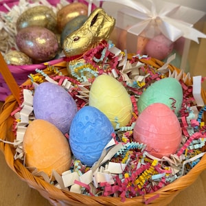 May include: A wicker basket filled with colorful pastel Easter eggs and a gold chocolate bunny. The basket is lined with shredded paper in pink, yellow, and white.