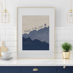 May include: Framed art print with the text "PROGRESS IS NOT LINEAR" above a silhouette of a person hiking on a mountain range. The artwork features shades of blue and beige, with a white border.