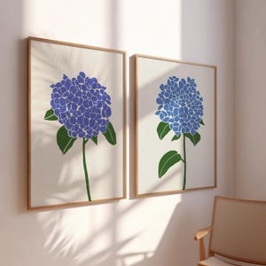 May include: Two framed art prints featuring stylized blue hydrangea flowers. Each print has a cream-colored background, a wooden frame, and a green stem and leaves. The flowers are a vibrant blue, creating a striking contrast.