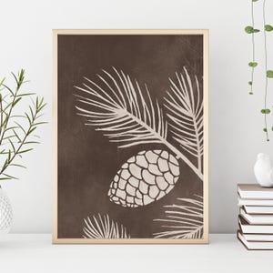 Może przedstawiać: Oprawiony wydruk artystyczny przedstawiający stylizowaną ilustrację szyszki i igieł sosnowych w kolorze kremowym na ciemnobrązowym tle. Grafika jest w złotej ramie i jest wyświetlana na białej powierzchni.