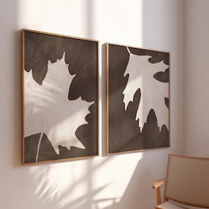 May include: Two framed wall art pieces. Each features a large white maple leaf silhouette against a dark brown background. The frames are a warm gold color, adding a touch of elegance to the minimalist design. The artwork is displayed on a white wall.