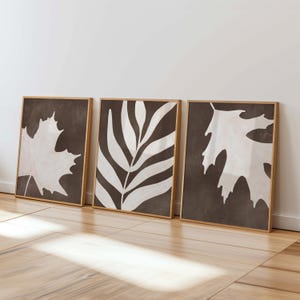 May include: Three framed art prints with botanical designs. The prints have a dark brown background with white leaf and plant illustrations. The frames are light brown. The artwork is leaning against a white wall.