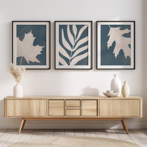 Peut inclure: Trois impressions botaniques encadrées avec des cadres noirs. Chaque impression présente un motif de feuille différent en beige sur un fond bleu canard foncé. Les impressions sont affichées au-dessus d'un meuble en bois clair avec un vase et des objets décoratifs.