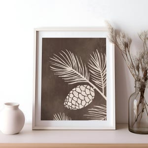 Może przedstawiać: Oprawiony wydruk z botaniczną ilustracją szyszki i gałęzi w kolorze kremowym na ciemnobrązowym tle. Grafika jest prezentowana w białej ramie, tworząc minimalistyczną estetykę. Obraz jest umieszczony na białej p&oacute;łce.