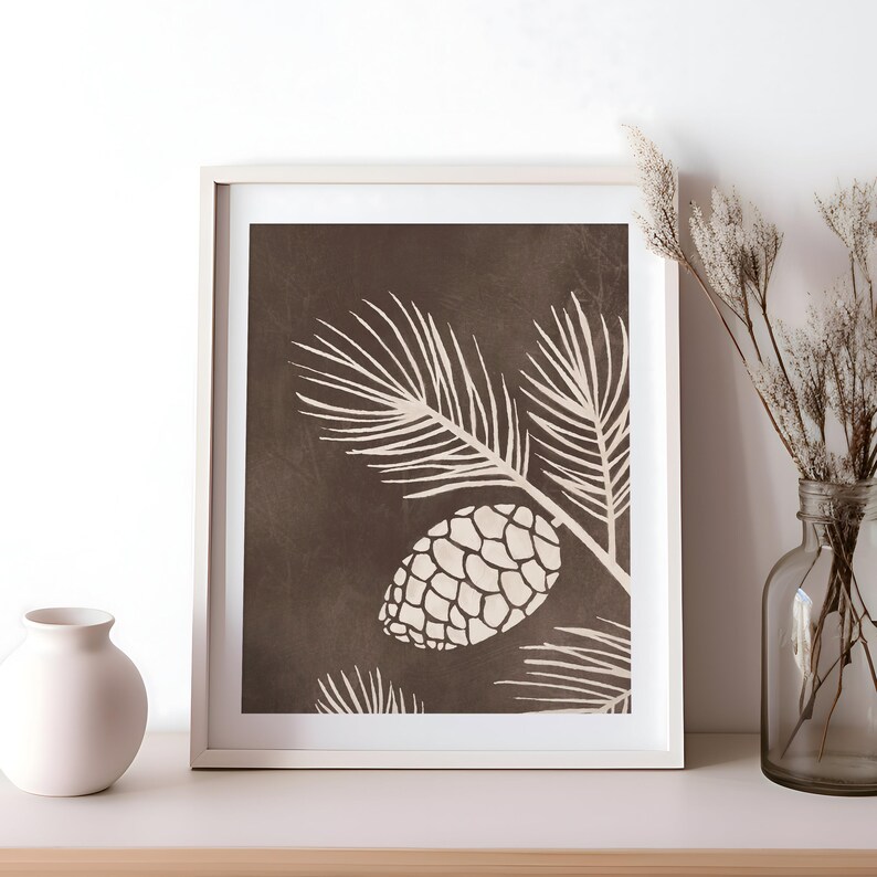 Może przedstawiać: Oprawiony wydruk z botaniczną ilustracją szyszki i gałęzi w kolorze kremowym na ciemnobrązowym tle. Grafika jest prezentowana w białej ramie, tworząc minimalistyczną estetykę. Obraz jest umieszczony na białej p&oacute;łce.