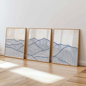 Może przedstawiać: Trzy oprawione wydruki artystyczne z minimalistycznym projektem krajobrazu górskiego. Każdy wydruk ma jasnobeżowe tło z niebieskimi falistymi liniami przedstawiającymi góry. Ramki wykonane są z jasnego drewna.