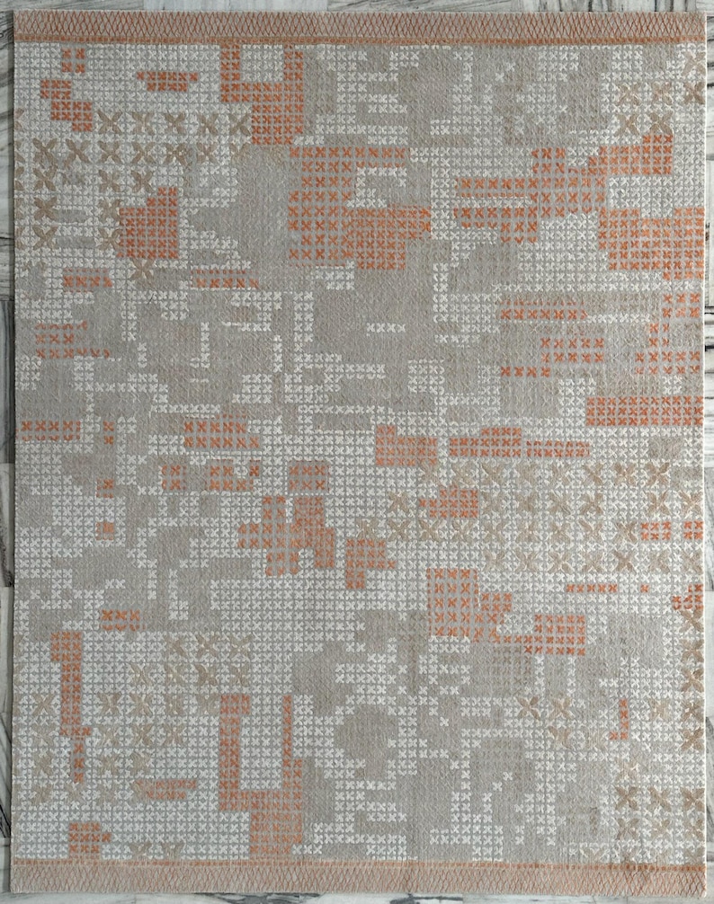 May include: A rectangular rug with a geometric pattern. The rug features a light gray background with a repeating pattern of small white and orange cross-stitch-like shapes. The border is a solid light orange color.