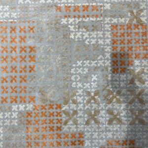 May include: Close-up of a textile with a repeating pattern of orange, white, and tan cross-stitch-like designs on a light blue background. The pattern is arranged in a grid-like fashion, creating a textured appearance.