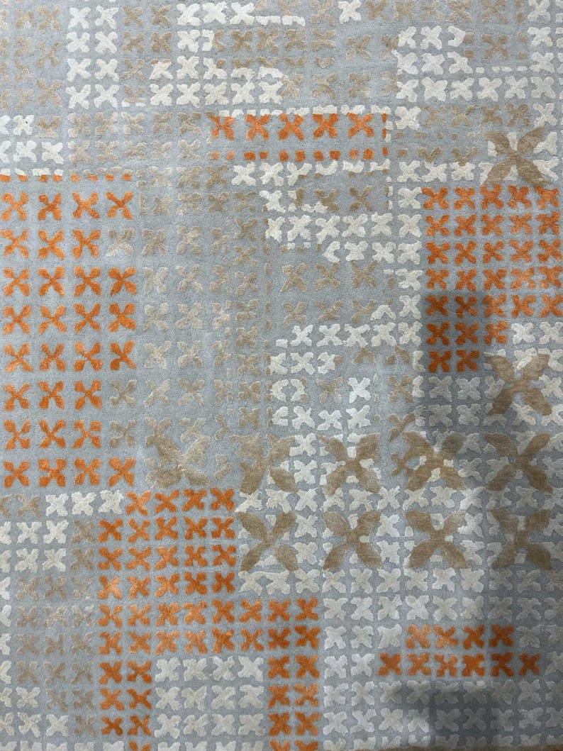 May include: Close-up of a textile with a repeating pattern of orange, white, and tan cross-stitch-like designs on a light blue background. The pattern is arranged in a grid-like fashion, creating a textured appearance.