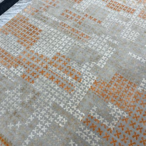 May include: A gray rug with a repeating pattern of orange and white cross-shaped designs. The rug has a geometric pattern with stripes of color and texture, creating a modern and stylish look. The rug is made of a soft material.