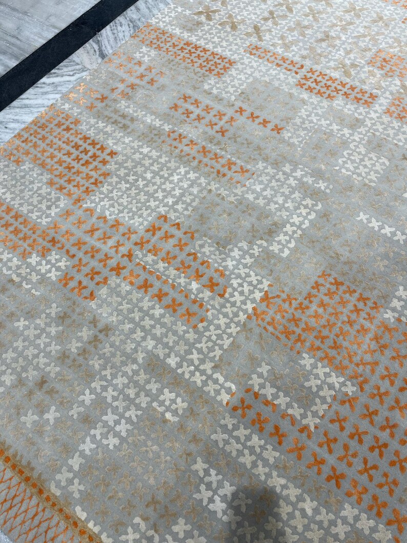 May include: A gray rug with a repeating pattern of orange and white cross-shaped designs. The rug has a geometric pattern with stripes of color and texture, creating a modern and stylish look. The rug is made of a soft material.