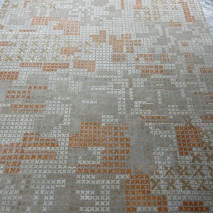 May include: A rectangular area rug with a geometric pattern. The rug features a light gray background with a repeating pattern of orange, white, and tan cross-stitch-like designs. The border is a darker orange with a woven texture.