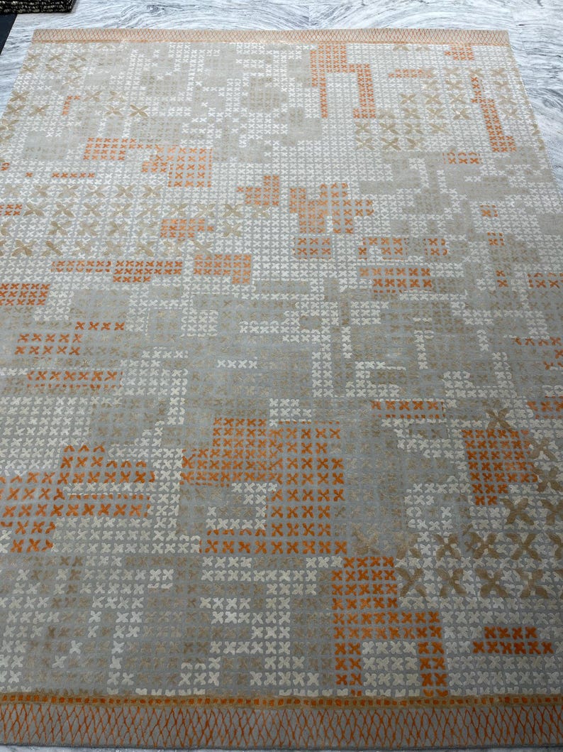 May include: A rectangular area rug with a geometric pattern. The rug features a light gray background with a repeating pattern of orange, white, and tan cross-stitch-like designs. The border is a darker orange with a woven texture.