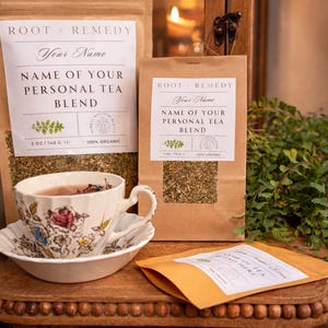 May include: An image of tea products, including a teacup and saucer with floral designs, a small wooden table, and several brown paper bags of tea. The bags have labels that read "ROOT + REMEDY" and "NAME OF YOUR PERSONAL TEA BLEND". The tea blend is 100% organic.