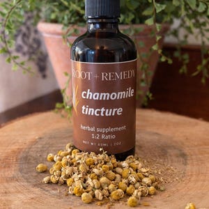 May include: A brown glass bottle of chamomile tincture with a black dropper, labeled "ROOT + REMEDY." The label reads "chamomile tincture, herbal supplement 1:2 Ratio, NET WT 60ML | 2oz." Dried chamomile flowers are scattered on a wooden surface.