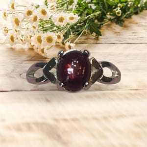 May include: A silver-toned ring featuring a large, oval, deep red gemstone. The ring has decorative accents on the sides and is set against a wooden background with white flowers.