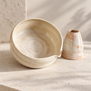 Può includere: Una ciotola in ceramica con un piccolo beccuccio e un contenitore conico abbinato, entrambi caratterizzati da uno smalto color crema con macchie marroni e sottili accenti rosso-bruni. La ciotola ha una forma arrotondata e il contenitore è affusolato.