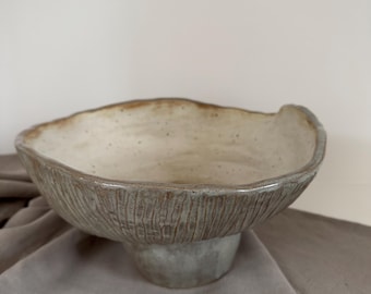 Puede incluir: Un cuenco de cerámica hecho a mano con un exterior texturizado y un interior de color claro. El cuenco tiene un borde ancho e irregular y se asienta sobre una base corta de pedestal. El color es un gris verdoso apagado con acentos marrones.
