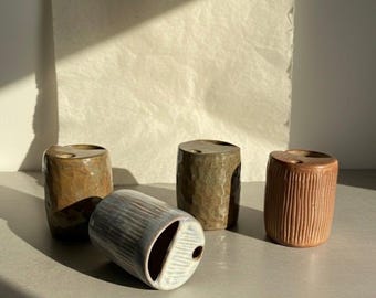 May include: Four cylindrical ceramic holders with varied surface textures and finishes. Three stand upright while one lies on its side. Each piece features a flat top with a circular opening and a larger semi-circular cutout, showcasing earthy tones.