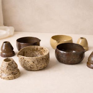 May include: A collection of ceramic pottery including three small bowls with pouring spouts and three conical-shaped objects. The items feature various earth-toned glazes in shades of brown, tan, and cream with speckled and smooth textures.