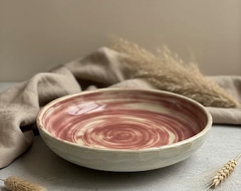 May include: A handmade ceramic bowl with a spiral design in shades of red and cream. The bowl has a wide, shallow shape and a textured, off-white exterior. The interior features a swirled pattern. Decorative wheat stalks are in the background.